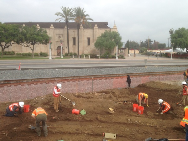 Workers excavating a historic water system in front of railroad tracks and the Mission San Gabriel