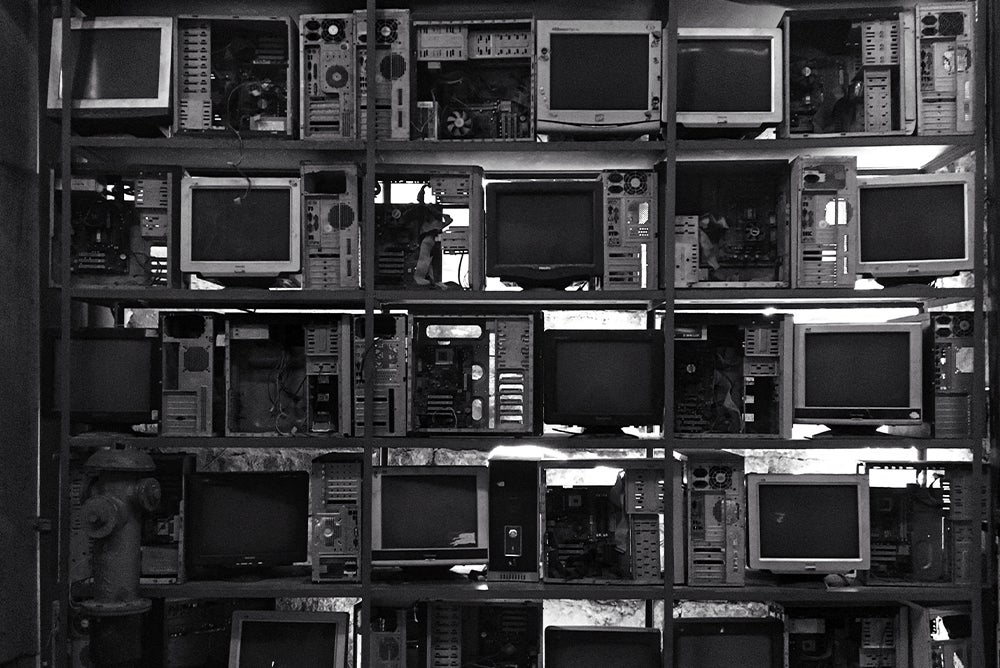Computer monitors, with the screen turned off, on row of shelves. Opened desktops are next to some of the monitors.