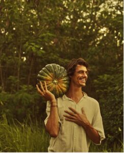 Evan Rilling smiling and look to the right. His left hand is placed on his chest. His right hand holds a large squash on his shoulder.