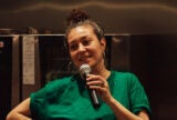 Zahara Gómez Lucini holds a microphone with mouth open in mid-speak. She wears a green t-shirt and stands in a kitchen, with an oven behind her.