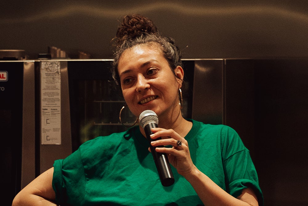 Zahara Gómez Lucini holds a microphone with mouth open in mid-speak. She wears a green t-shirt and stands in a kitchen, with an oven behind her.