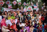 A tight crowd of men and women, some sitting down, some standing. Political banners and pictures are seen above the people.
