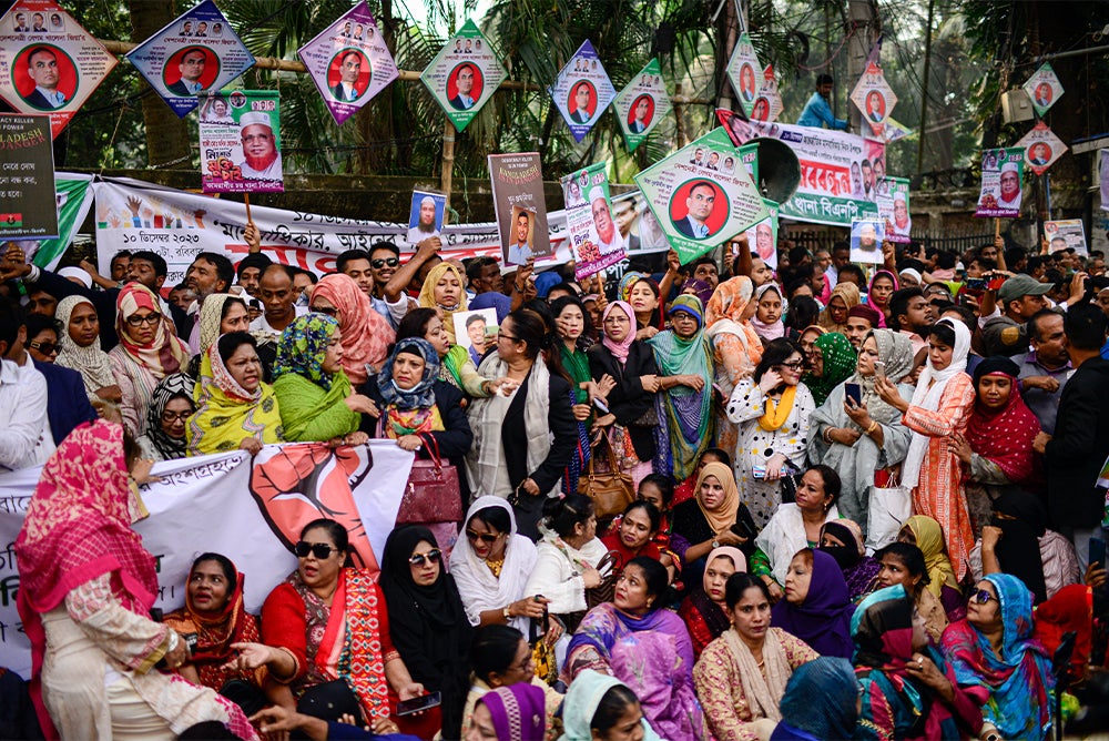 A tight crowd of men and women, some sitting down, some standing. Political banners and pictures are seen above the people.