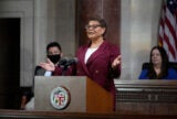Karen Bass stands in front of a podium with both her hands raised in an open gesture at L.A. City Hall. Two council members are sitting and are visible in the background.