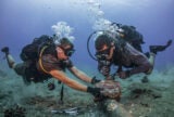 Two men in diver's equipment in the ocean focused on removing corroded zinc anodes from an undersea cable.