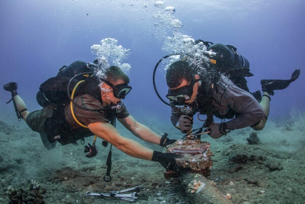 Two men in diver's equipment in the ocean focused on removing corroded zinc anodes from an undersea cable.