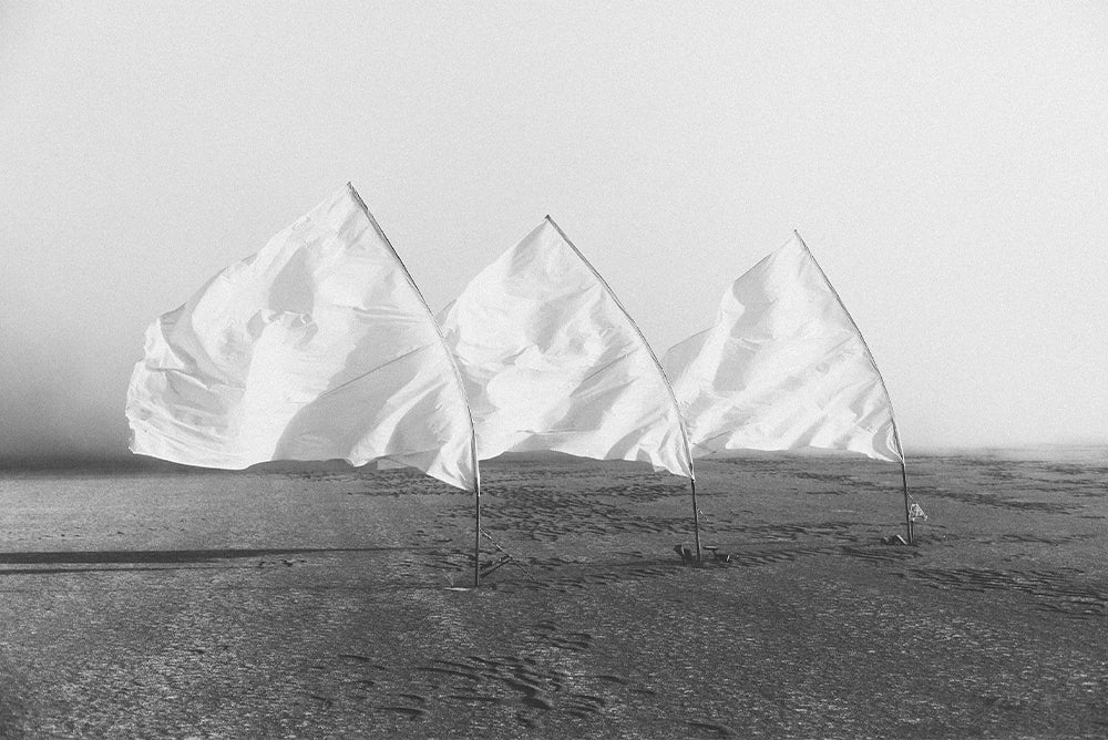Black and white photo of three white flags and their poles strung to a dirt empty ground. The flags are flying to the left, seemingly from wind, with the creases making the flags look like icebergs.