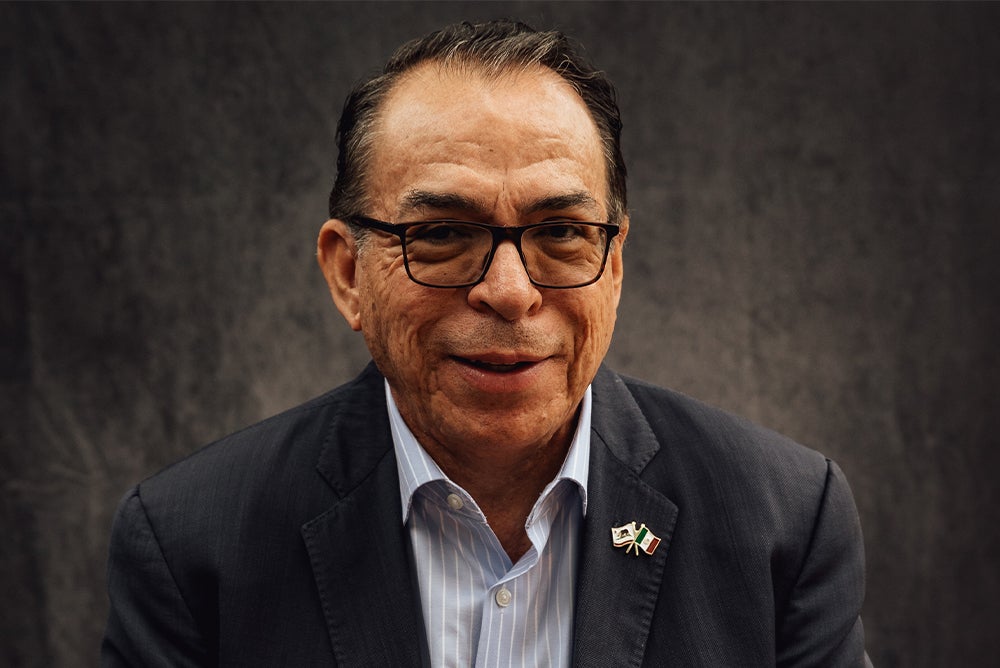 A headshot of Alfredo Corchado smiling against a gray-black backdrop. He wears black-rimmed glasses and a black jacket on top of a blue collared shirt. A small pin of California's official state flag and Mexico's official flag waving is pinned to right side of his jacket.