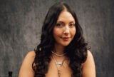 A headshot of Anita Herrera smiling against a gray-black backdrop. Her face is slightly turned to the right. A pearl necklace is around her neck. She also wears a necklace that says, "Anita."